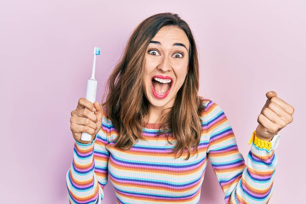 Young hispanic girl holding electric toothbrush screaming proud, celebrating victory and success very excited with raised arms  - Φωτογραφία, εικόνα