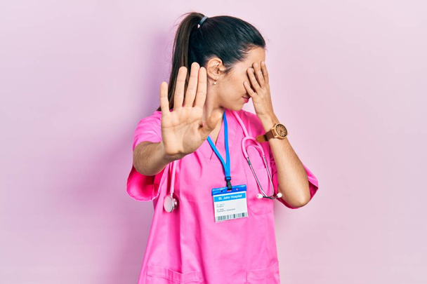 Young brunette woman wearing doctor uniform and stethoscope covering eyes with hands and doing stop gesture with sad and fear expression. embarrassed and negative concept.  - Photo, Image