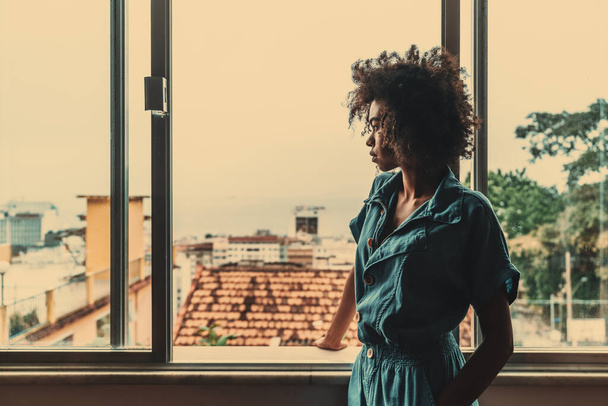 Eine nachdenkliche junge schwarze Frau in blauen Overalls und mit lockigem afrikanischen Haar steht drinnen am Fenster ihres Hauses und blickt nachdenklich nach draußen, links ist ein Kopierplatz. - Foto, Bild