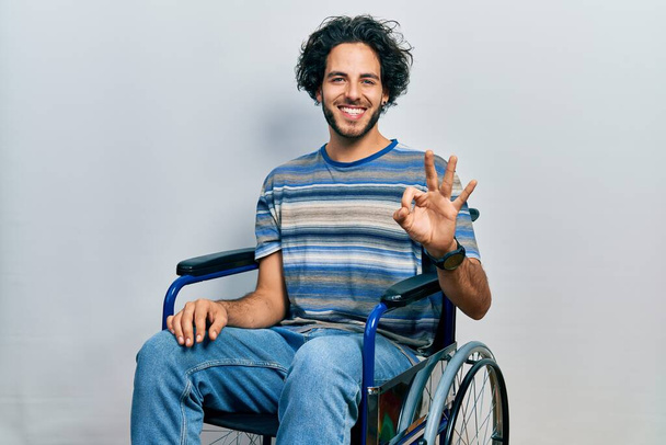 Jóképű spanyol férfi, aki tolószéken ül, mutatva és mutatva a hármas számú ujjaival, miközben magabiztosan és boldogan mosolyog..  - Fotó, kép