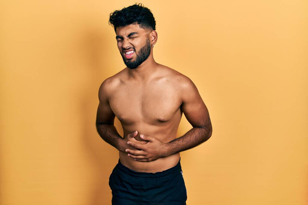 Arab man with beard wearing swimwear shirtless with hand on stomach because nausea, painful disease feeling unwell. ache concept.  - Foto, Imagen