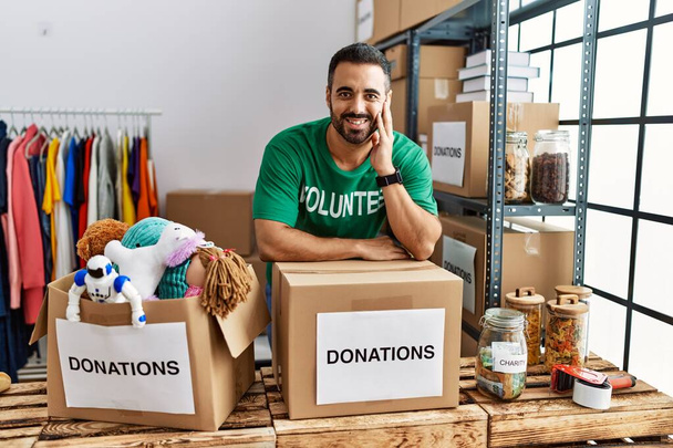 Young hispanic man wearing volunteer uniform leaning on donations box at charity center - Photo, Image