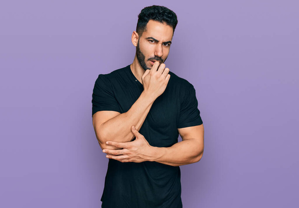 Hispanic man with beard wearing casual black t shirt looking stressed and nervous with hands on mouth biting nails. anxiety problem.  - Foto, afbeelding