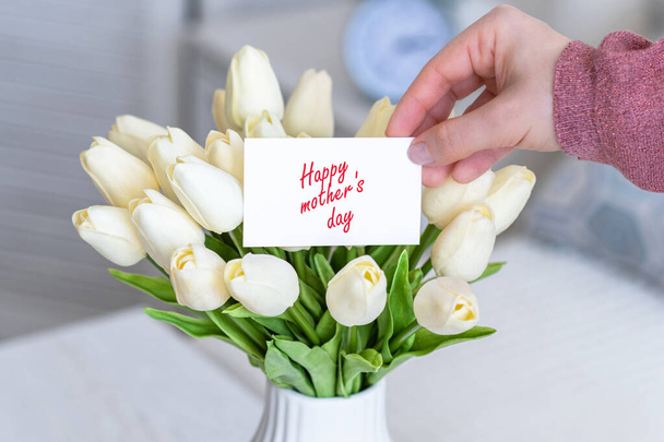 Woman pulling greeting card with Happy mother's day words from bouquet of white tulips flowers in a white vase on the table.  - Фото, изображение