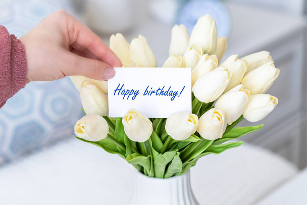 Woman pulling greeting card with Happy birthday words from bouquet of white tulips flowers in a white vase on the table.  - Foto, immagini