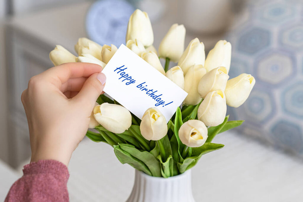 Woman pulling greeting card with Happy birthday words from bouquet of white tulips flowers in a white vase on the table.  - Foto, Imagem