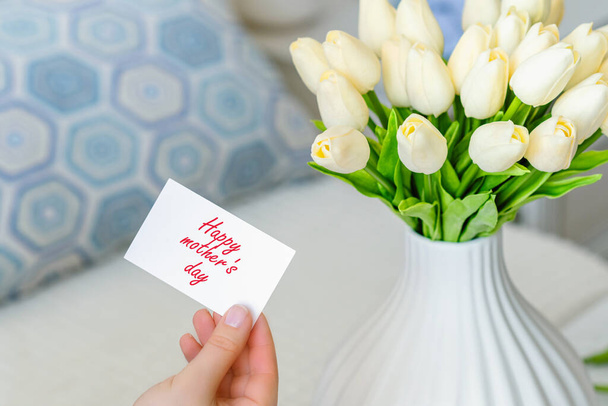 Woman pulling greeting card with Happy mother's day words from bouquet of white tulips flowers in a white vase on the table.  - Fotografie, Obrázek