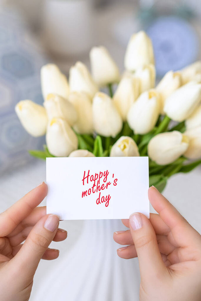 Woman pulling greeting card with Happy mother's day words from bouquet of white tulips flowers in a white vase on the table.  - 写真・画像