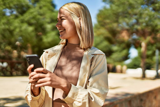 Jeune fille blonde souriant heureux en utilisant un smartphone à la ville. - Photo, image
