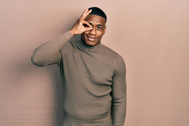 Young black man wearing casual turtleneck sweater doing ok gesture with hand smiling, eye looking through fingers with happy face.  - Photo, Image
