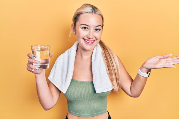 Giovane ragazza bionda che indossa abbigliamento sportivo bere bicchiere d'acqua celebrando il successo con sorriso felice e l'espressione del vincitore con la mano alzata  - Foto, immagini