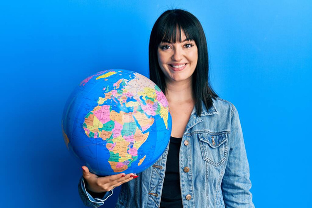 Молода іспаномовна жінка тримає світовий м'яч позитивно і щасливо стоїть і посміхається з впевненою посмішкою, показуючи зуби
  - Фото, зображення