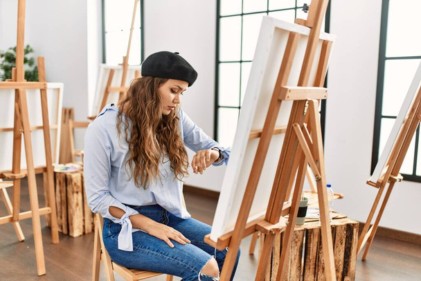 Young hispanic artist woman painting on canvas at art studio checking the time on wrist watch, relaxed and confident  - Photo, Image