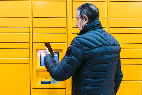 Mężczyzna w szarym płaszczu patrzy na komórkę, żeby odebrać paczkę z szafki. Koncepcja wiadomości, porównaj online, e-commerce i pakiety - Zdjęcie, obraz