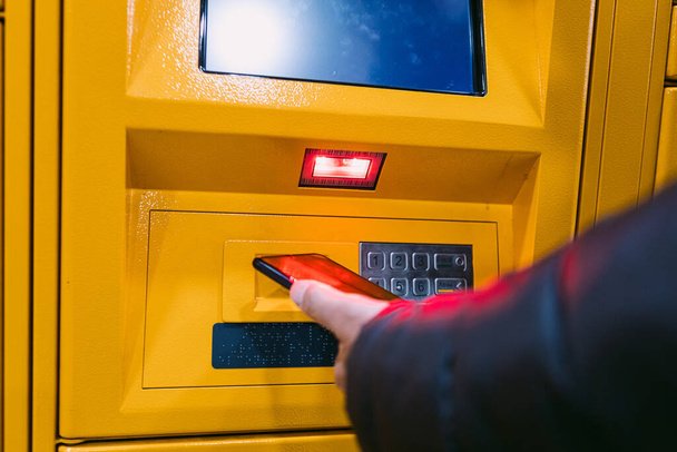 Detail of the arm of a man scanning a code on the mobile phone to pick up a package from the yellow locker. Messaging concept, compare online, e-commerce and packages - Photo, Image