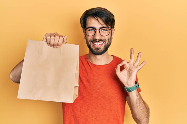 Young hispanic man holding paper bag with bread doing ok sign with fingers, smiling friendly gesturing excellent symbol  - Foto, afbeelding