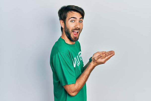 Молодой латиноамериканец в футболке волонтера, указывая в сторону с открытыми руками ладонями, показывая пространство для копирования, представляя рекламу улыбающейся счастливой  - Фото, изображение