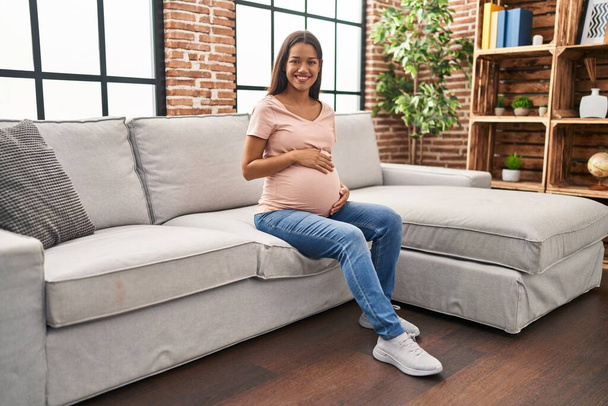 Mujer latina joven embarazada sonriendo confiada sentada en el sofá en casa - Foto, imagen