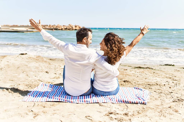 Střední věk hispánský pár na zadní pohled sedí na ručníku objímání na pláži. - Fotografie, Obrázek