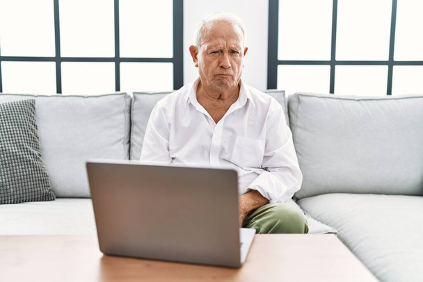 Ανώτερος άνθρωπος που χρησιμοποιεί φορητό υπολογιστή στο σπίτι κάθεται στον καναπέ σκεπτικός και νευρικός, συνοφρυωμένος αναστατωμένος λόγω του προβλήματος. αρνητικό πρόσωπο.  - Φωτογραφία, εικόνα