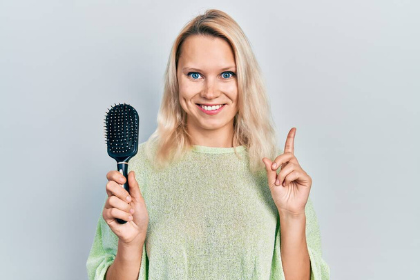 Beautiful caucasian blonde woman styling hair using comb smiling happy pointing with hand and finger to the side  - Φωτογραφία, εικόνα