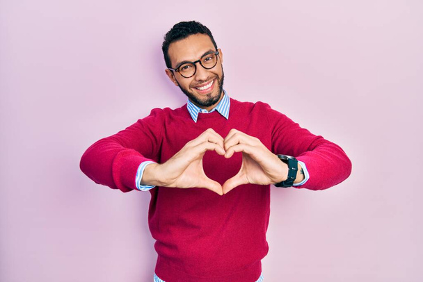 Hombre hispano con barba vistiendo camisa de negocios y gafas sonriendo en amor haciendo forma de símbolo del corazón con las manos. concepto romántico.  - Foto, Imagen