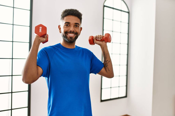 Handsome hispanic man doing exercise training at the gym holding dumbbells - Fotoğraf, Görsel