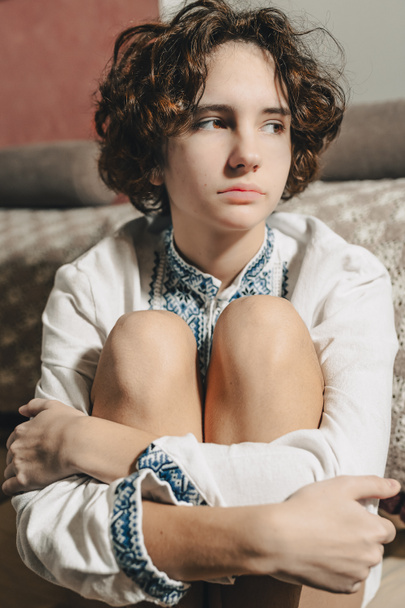 sad young woman with brown curly hair tied in bun, wearing white ethnic embroidered shirt, sits in depressed pose, bending legs, clasped hands - Foto, Bild