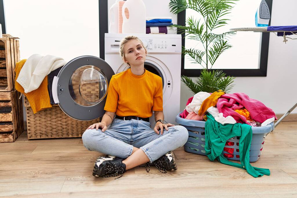 Jeune femme blonde faisant la lessive assis à côté de la machine à laver l'air somnolent et fatigué, épuisé par la fatigue et la gueule de bois, les yeux paresseux le matin.  - Photo, image