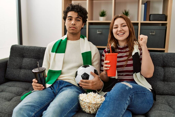 Young hooligan couple supporting soccer team sitting on the sofa at home. - Zdjęcie, obraz