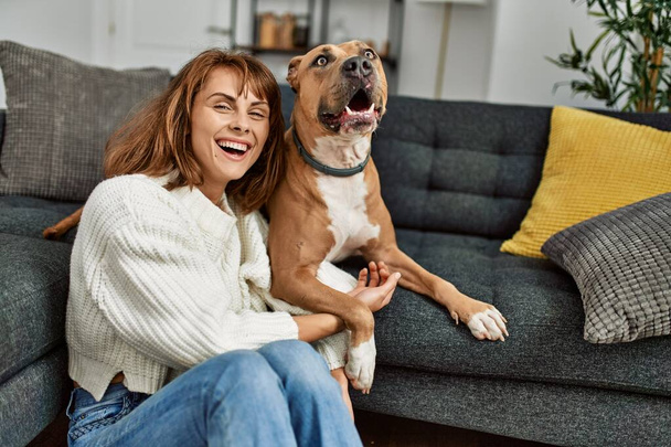 Молодая белая женщина улыбается уверенно обнимая собаку сидя дома на полу - Фото, изображение
