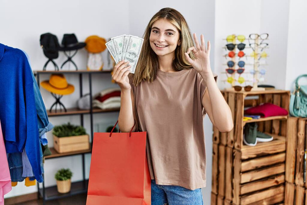 Jonge blonde vrouw met boodschappentassen en dollar in de kledingwinkel doet ok teken met de vingers, glimlachend vriendelijk gebaar uitstekend symbool  - Foto, afbeelding