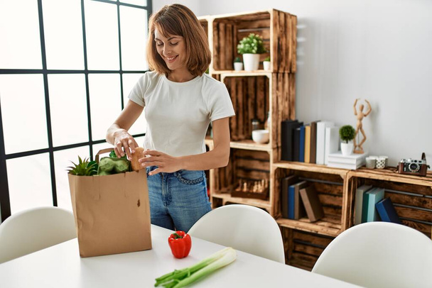 Young caucasian woman holding food of groceries paper bag at home - Foto, Imagen