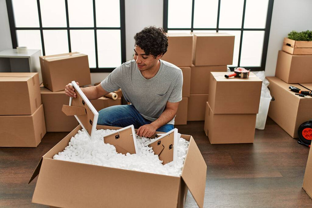 Joven hombre hispano sonriendo feliz unboxing caja de cartón en nuevo hogar. - Foto, Imagen