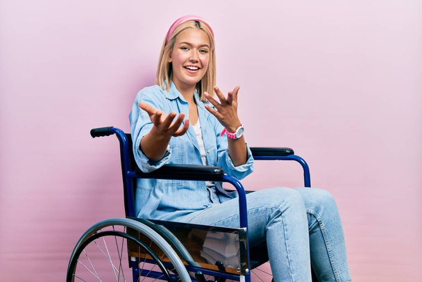 Hermosa mujer rubia sentada en silla de ruedas sonriente alegre ofreciendo manos dando asistencia y aceptación.  - Foto, imagen