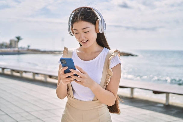 Joven chica china sonriendo feliz usando auriculares y teléfono inteligente en el paseo marítimo. - Foto, Imagen
