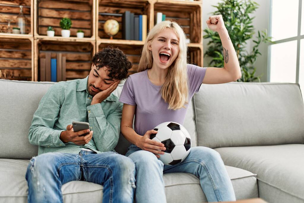Woman smiling happy watching soccer match and boyfriend boring using smartphone at home. - 写真・画像