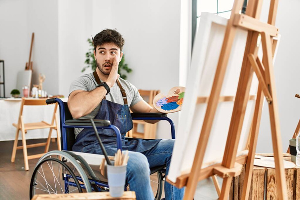 Young hispanic man sitting on wheelchair painting at art studio hand on mouth telling secret rumor, whispering malicious talk conversation  - Foto, Imagem