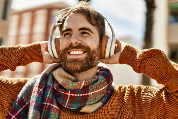 Mladý kavkazský muž s plnovousem poslouchající hudbu nošení sluchátek venku za slunečného dne - Fotografie, Obrázek