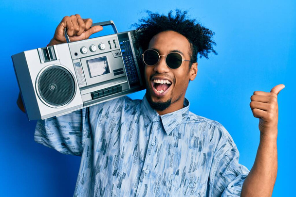 Young african american man with beard holding boombox, listening to music pointing thumb up to the side smiling happy with open mouth  - Photo, Image