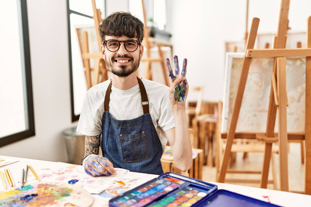 Spanyol férfi szakállal a művészeti stúdióban, mutatva és mutatva a hármas számú ujjal, miközben magabiztosan és boldogan mosolyog..  - Fotó, kép