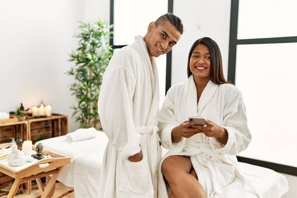 Junges lateinisches Paar im Bademantel mit Smartphone im Schönheitszentrum. - Foto, Bild