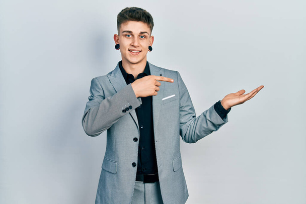 Junge kaukasische Junge mit Ohren Erweiterung in Business-Jacke erstaunt und lächelnd in die Kamera, während mit der Hand präsentieren und mit dem Finger zeigen.  - Foto, Bild