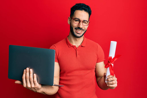 Junger hispanischer Mann mit Laptop und Diplom, der augenzwinkernd in die Kamera blickt, mit sexy Gesichtsausdruck, fröhlichem und glücklichem Gesicht.  - Foto, Bild