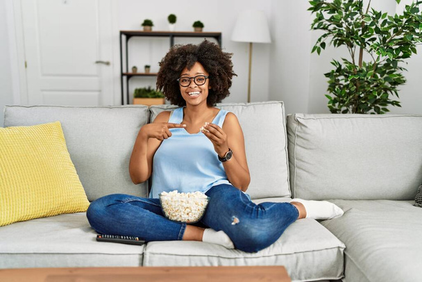 Afrikai amerikai nő afro haj ül a kanapén eszik popcorn otthon mosolyogva boldog rámutatva a kezét és ujját  - Fotó, kép