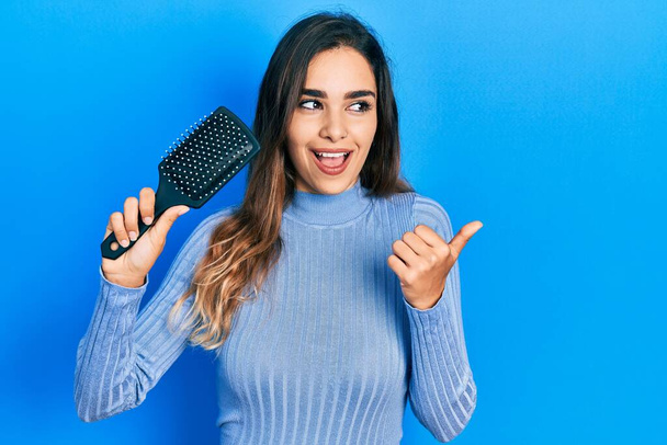 Young hispanic girl holding hairbrush pointing thumb up to the side smiling happy with open mouth  - Photo, Image