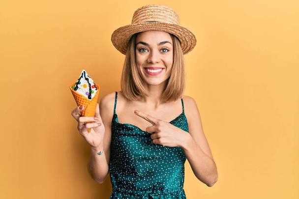 Joven mujer rubia caucásica comiendo helado sonriendo feliz señalando con la mano y el dedo  - Foto, imagen