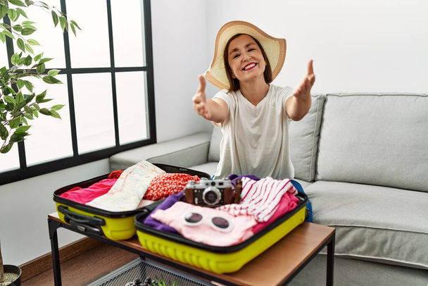 Kaunis keski-ikäinen latino nainen pakkaa kesävaatteita matkalaukussa katsoen kameraa hymyillen avosylin halausta varten. iloinen ilme käsittää onnellisuuden.  - Valokuva, kuva