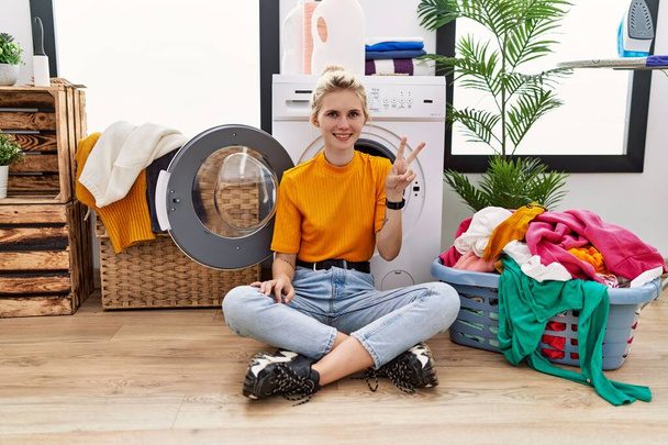 jonge blonde vrouw doen wasgoed zitten door wasmachine tonen en wijzen met vingers nummer twee terwijl glimlachen zelfverzekerd en gelukkig.  - Foto, afbeelding