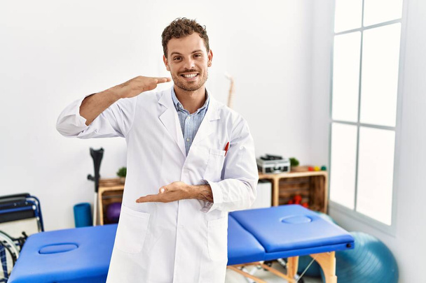 Handsome young man working at pain recovery clinic gesturing with hands showing big and large size sign, measure symbol. smiling looking at the camera. measuring concept.  - Fotó, kép